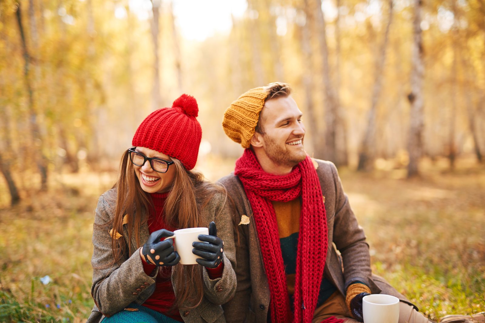 Smiling couple in warm hats and scarves sitting in an autumn forest holding hot drinks