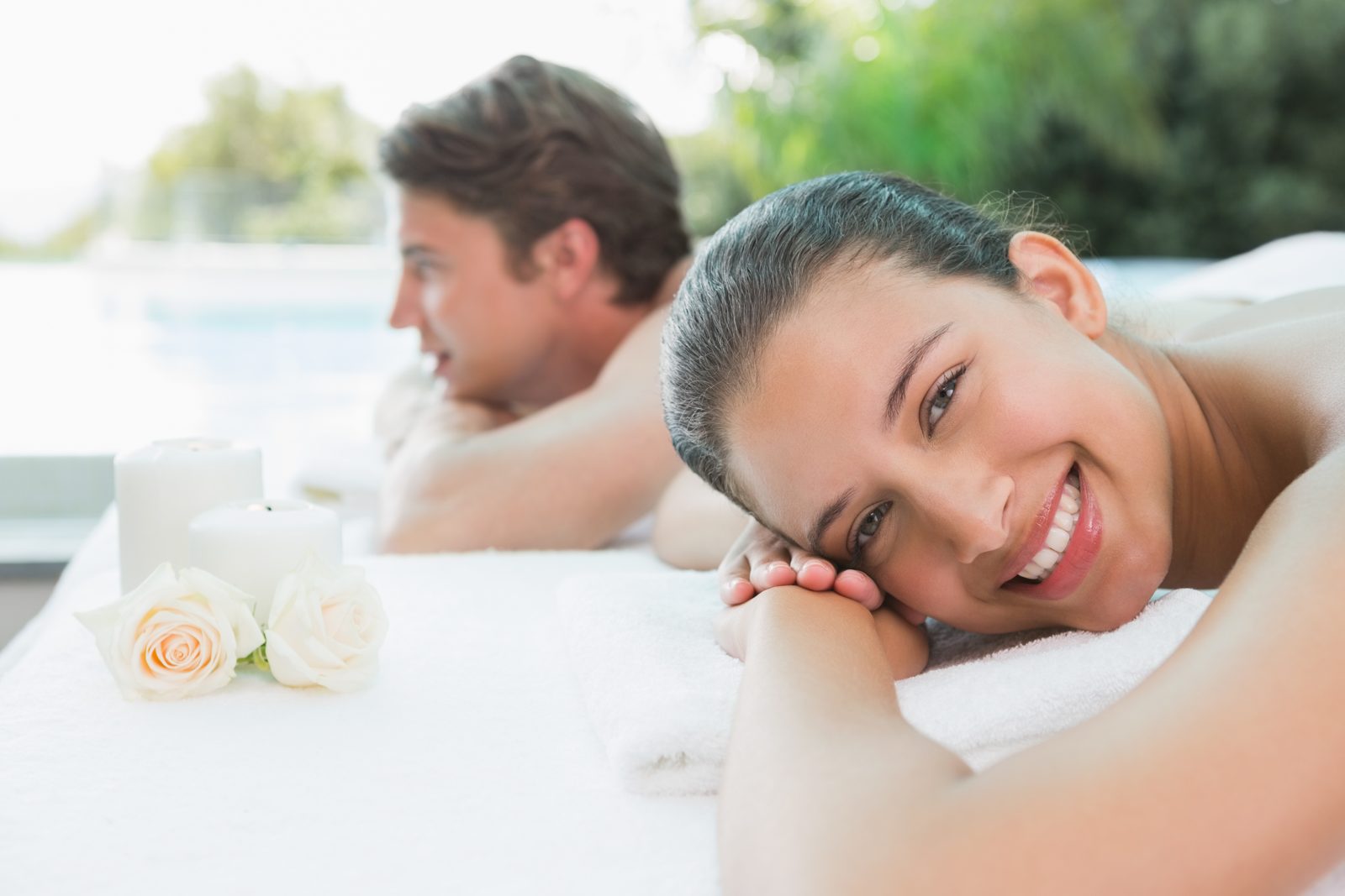 Smiling woman and man lying side by side on massage tables with candles and white roses